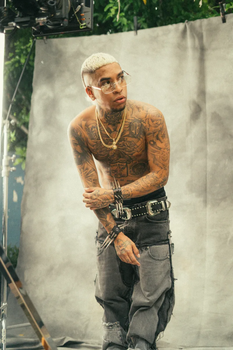 Artista con el torso tatuado, pelo platinado y cadenas de oro, frente a un lienzo gris de estudio con humo en el aire
